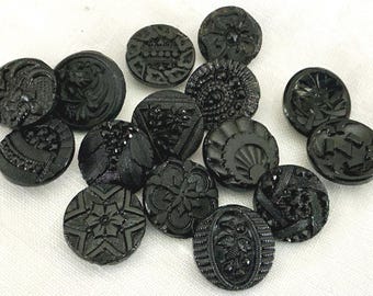 May include: A collection of black vintage buttons with various embossed designs. The buttons are round and feature floral, geometric, and abstract patterns. They are arranged on a white surface.