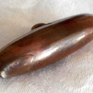 May include: A polished, oval-shaped, bronze-colored metal object with a small, circular attachment on top. The object has a smooth, reflective surface and appears to be a decorative element or hardware component.