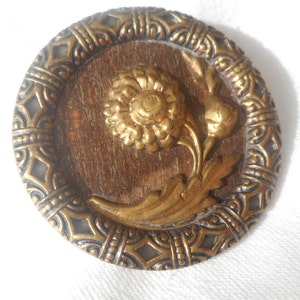 May include: A round, gold-toned button with a raised, ornate design. The center features a stylized flower with leaves, all in a gold finish. The button has a decorative border with a repeating pattern.