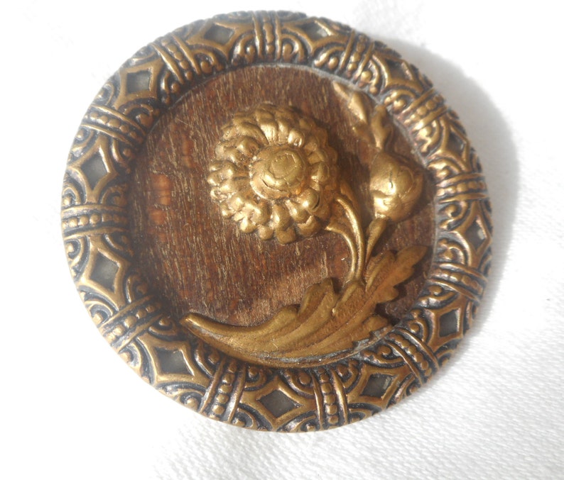 May include: A round, gold-toned button with a raised, ornate design. The center features a stylized flower with leaves, all in a gold finish. The button has a decorative border with a repeating pattern.
