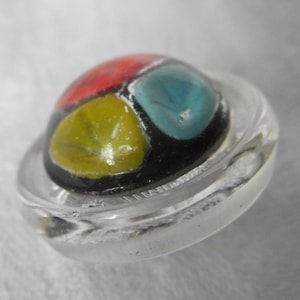 May include: A colorful glass button with red, yellow, blue, and black enamel. The button is set in a clear plastic base.