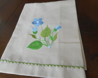 May include: A white cotton tea towel with a green embroidered border and a blue and green floral design.
