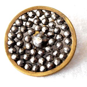 May include: A round, antique button with a gold-colored metal frame. The button's center features a large, dark gray, faceted bead surrounded by smaller, similar beads. The button has a vintage aesthetic and is likely from a garment.