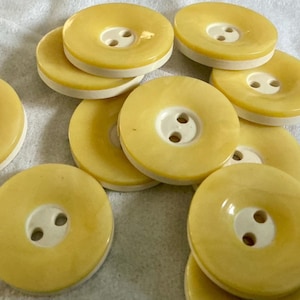 May include: A collection of round, yellow buttons with a white center and two holes. The buttons are scattered on a white surface. The buttons have a glossy finish and a slightly raised edge.