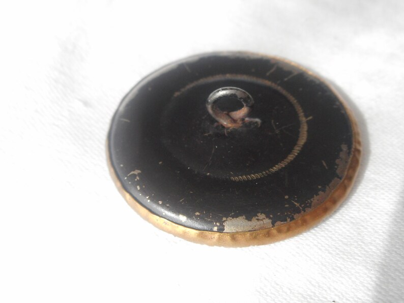 May include: A black button with a gold rim and a metal loop on the top.