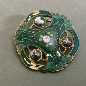 May include: Vintage round brooch with a green enamel floral design and gold-tone metal accents. The brooch features three clear, faceted stones and a central painted pink rose. The edges show signs of wear, adding to its antique charm.
