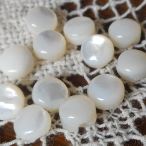 May include: A collection of white, round, pearl-like buttons. The buttons are arranged on a white crocheted surface.