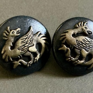 May include: Two round, dark gray buttons with a raised, gold-colored mythical creature design. The creature has a bird-like head, wings, and a serpentine tail. The buttons are likely for clothing or crafts.