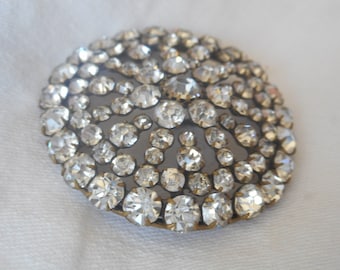 May include: A round, silver-toned brooch with a design of many small, clear rhinestones set in a circular pattern.
