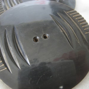 May include: A black plastic button with a decorative, carved design. The button has two holes for sewing.