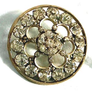May include: A round, vintage button with a gold-tone metal frame. The button is adorned with numerous clear, sparkling rhinestones arranged in a floral pattern. The design features a central cluster and surrounding circular accents.