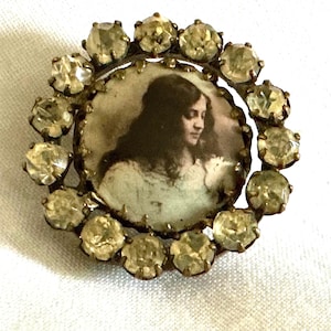 May include: A circular brooch featuring a central sepia-toned portrait of a person with long dark hair wearing a light-colored garment. The portrait is surrounded by a border of clear, faceted, round-cut stones set in a metallic frame.