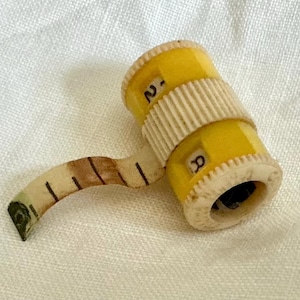 May include: A vintage yellow and cream-colored miniature tape measure. The tape measure is cylindrical with a cream-colored ribbed band and a pull-out tape marked with black lines and numbers. The numbers are visible on the yellow cylinder.