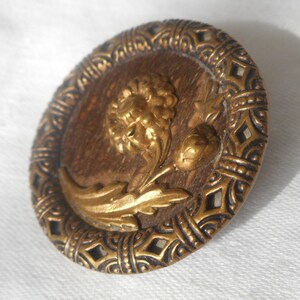 May include: A round, gold-toned button with a raised, ornate design. The center of the button features a stylized flower with leaves, all in gold. The outer edge of the button is a decorative, black-and-gold border.