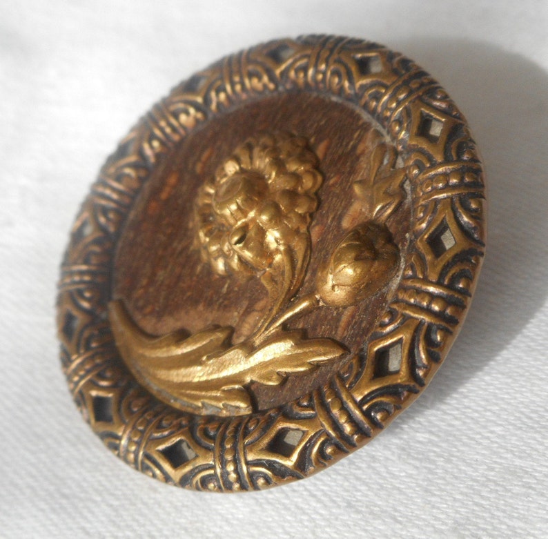 May include: A round, gold-toned button with a raised, ornate design. The center of the button features a stylized flower with leaves, all in gold. The outer edge of the button is a decorative, black-and-gold border.