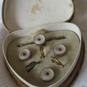 May include: A heart-shaped box with a velvet lining contains a set of cufflinks and a tie bar. The cufflinks and tie bar are made of gold metal and have white mother-of-pearl settings with red gemstones in the center.