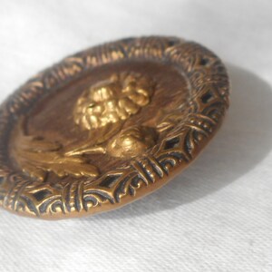 May include: A gold-toned metal button with a raised design of a flower and leaves. The button has a decorative, intricate border.