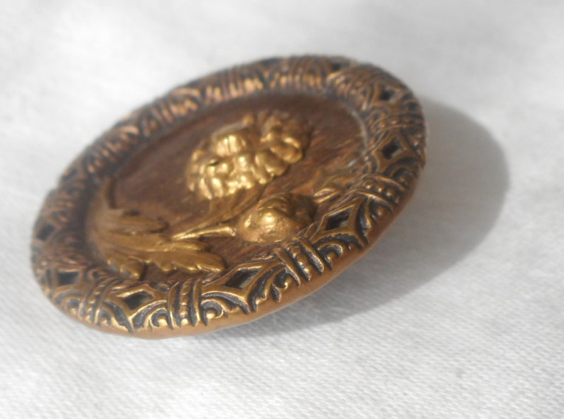 May include: A gold-toned metal button with a raised design of a flower and leaves. The button has a decorative, intricate border.
