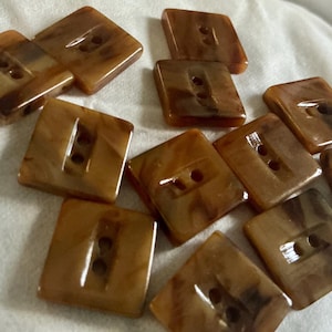 May include: A collection of square, brown buttons with two holes each. The buttons have a marbled appearance with darker brown streaks, and are arranged on a white surface. These buttons are suitable for sewing projects.