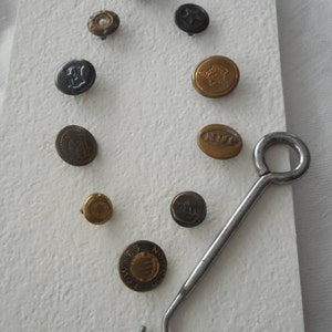 May include: A collection of vintage buttons in various colors and designs, including black, gold, and bronze. Some buttons feature embossed details, letters, or symbols. A metal button hook is also present.