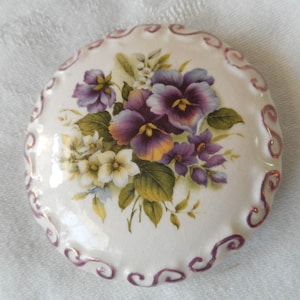May include: A white ceramic trinket box with a floral design. The box is round and has a raised edge with a gold and purple swirl pattern. The floral design features purple and yellow pansies with white flowers and green leaves.