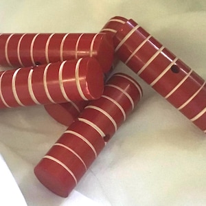 May include: A collection of four cylindrical wooden toggles featuring a deep red finish with evenly spaced, thin horizontal white stripes. Each toggle has a small, circular hole drilled through the center of the body for attachment purposes.