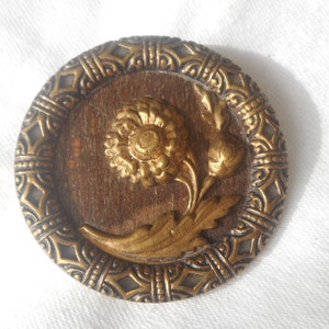 May include: A gold-toned metal button with a raised floral design. The button features a stylized flower with a central bud and leaves, all set against a wood-grain background. The button is surrounded by a decorative border.