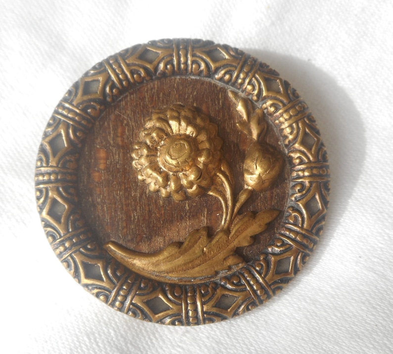 May include: A gold-toned metal button with a raised floral design. The button features a stylized flower with a central bud and leaves, all set against a wood-grain background. The button is surrounded by a decorative border.