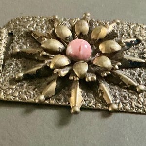 May include: A vintage gold-toned metal bracelet with a textured surface. Each of the three rectangular links features a central pink stone surrounded by gold-colored petal-like shapes. The bracelet is designed with a floral motif.