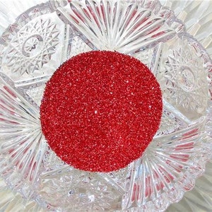 May include: A clear, cut-glass bowl with a circular mound of red glitter. The bowl has a starburst design with intricate patterns. The red glitter is densely packed, creating a vibrant contrast against the clear glass.