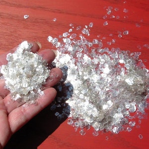 May include: A close-up of a hand holding a pile of iridescent, silver mica flakes. The flakes are scattered on a red surface.