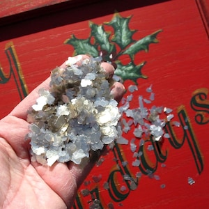 May include: A hand holding a pile of silver and gray glitter. The glitter is scattered on a red surface with the word "Holidays" written in black.