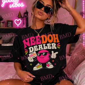 Może przedstawiać: Czarny t-shirt z napisem "NEEDOH DEALER" w kolorze różowym i pomarańczowym, z postacią z kreskówki w okularach przeciwsłonecznych. Postać to różowe koło z ramionami i nogami, trzymające znak z napisem "OFFICIAL NEEDOH DEALER".