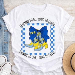 May include: White t-shirt featuring a blue bow and yellow cowboy boots with floral accents inside a blue and white checkered square. The text reads "LEARNING TO DO, DOING TO LEARN, EARNING TO LIVE, LIVING TO SERVE." The letters "FFA" are on the bow.