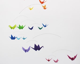 May include: A colorful origami crane mobile featuring multiple paper cranes in a variety of colors, including red, orange, yellow, green, blue, and purple. The cranes are suspended by thin wires against a white background.