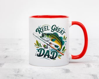 May include: White ceramic mug with a red interior and handle. The mug features a colourful graphic of a fish and fishing rod, with the text "Reel Great Dad" in teal and white. The design is set against a white background.