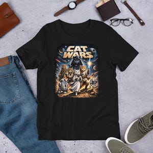 Może przedstawiać: Czarny t-shirt z grafiką "Cat Wars". Projekt przedstawia parodię postaci z Gwiezdnych Wojen jako koty, na tle kosmicznym. Koszulka wykonana jest z miękkiego materiału i nadaje się do noszenia na co dzień.