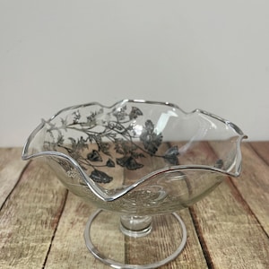 Op de afbeelding: Een heldere glazen voetkom met een zilveren rand en bloemenmotief. De kom heeft een geschulpte rand en staat op een cirkelvormige voet. Het bloemenmotief is zilverkleurig en bedekt de binnenkant van de kom.