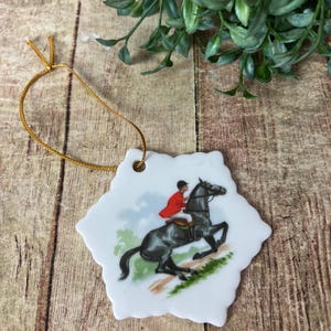 May include: White ceramic ornament with a scalloped edge, featuring a rider in a red coat on a black horse. The horse is jumping over a small hill. A gold string is attached for hanging.