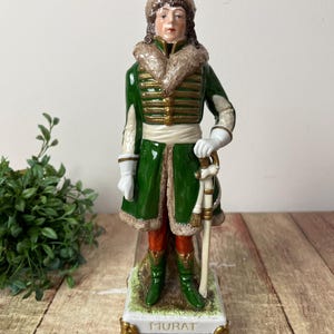 Puede incluir: Figura de porcelana de un personaje militar con abrigo verde con adornos dorados, guantes blancos y pantalones rojos. La figura lleva un sombrero con ribete de piel y sostiene una espada. La base está inscrita con el nombre "MURAT".