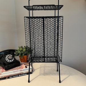 May include: A black metal magazine rack featuring a tall, rectangular frame with a perforated circular pattern on the sides and a flat top shelf. The structure is supported by four thin legs with protective caps at the base, providing a stable stand.
