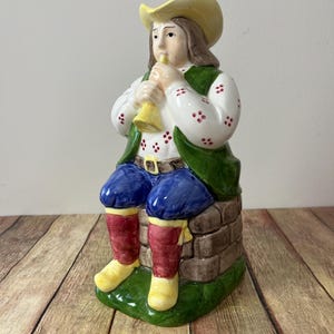 Könnte beinhalten: Eine Keramikfigur eines Menschen, der eine grüne Weste, eine blaue Hose, rote Stiefel und einen gelben Hut trägt und ein gelbes Musikinstrument spielt. Die Figur sitzt auf einem Steinsockel mit grünem Gras.