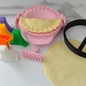 Puede incluir: Un molde rosa para hacer empanadillas con una empanadilla rellena en su interior, junto con un cortador circular negro, un c&iacute;rculo de masa y sellos de colores. Los sellos son naranja, verde, morado, amarillo y blanco. El sello naranja dice "MAH".