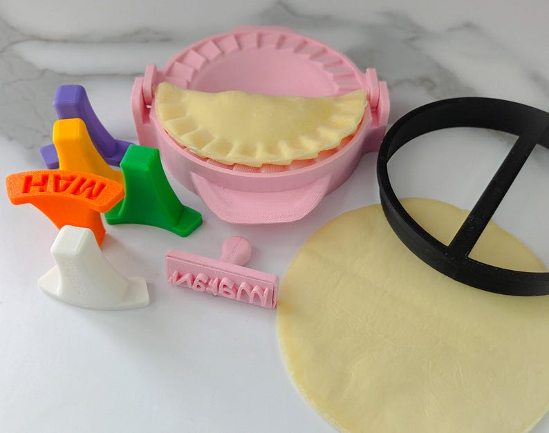 Puede incluir: Un molde rosa para hacer empanadillas con una empanadilla rellena en su interior, junto con un cortador circular negro, un c&iacute;rculo de masa y sellos de colores. Los sellos son naranja, verde, morado, amarillo y blanco. El sello naranja dice "MAH".