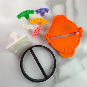 Puede incluir: Un surtido de coloridas herramientas de cocina, que incluyen una m&aacute;quina para hacer raviolis de color naranja brillante, un cortador circular negro y varios sellos con texto. Los sellos son de varios colores y dicen "APPLE", "CHEESE", "PIZZA" y "BOENE".