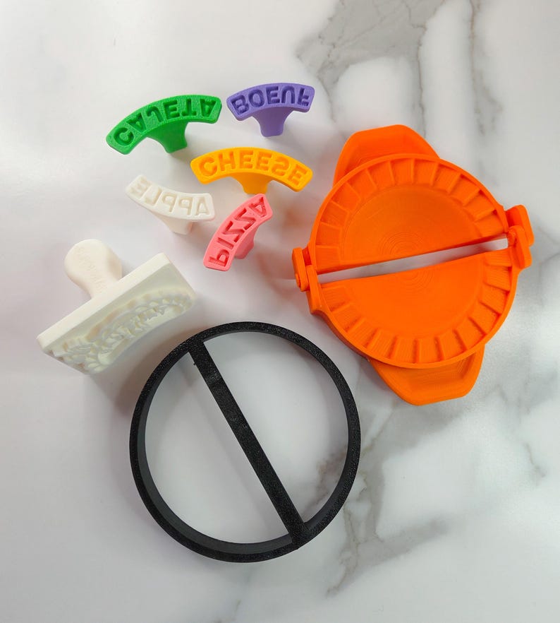 Puede incluir: Un surtido de coloridas herramientas de cocina, que incluyen una m&aacute;quina para hacer raviolis de color naranja brillante, un cortador circular negro y varios sellos con texto. Los sellos son de varios colores y dicen "APPLE", "CHEESE", "PIZZA" y "BOENE".