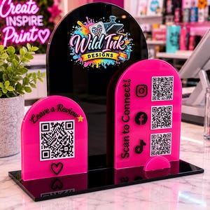 Pode incluir: Um expositor de acrílico preto e rosa choque com três painéis arqueados. O painel central preto exibe o logótipo Wild Ink Designs. Os dois painéis cor-de-rosa apresentam códigos QR e o texto Leave a Review e Scan to Connect.