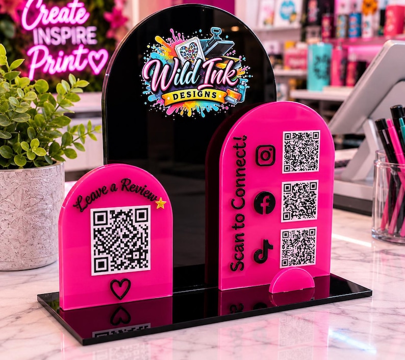 Op de afbeelding: Een zwarte en felroze acryl displaystandaard met drie boogvormige panelen. Het centrale zwarte paneel toont het Wild Ink Designs logo. De twee roze panelen bevatten QR-codes en de tekst Leave a Review en Scan to Connect.