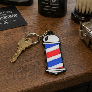 May include: A metal key attached to a rubberized keychain shaped like a traditional barber pole with diagonal red, white, and blue stripes. The keychain features a rounded white top and bottom, a black border, and a silver-toned metal split ring.