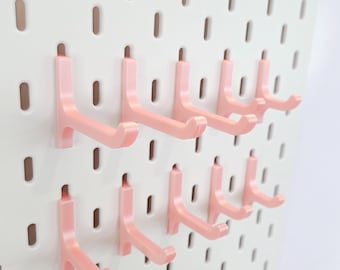 May include: A white pegboard with multiple pink plastic hooks. The hooks are in two rows and are designed to hang items on the pegboard.